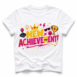 May include: A white short-sleeve t-shirt featuring a colorful pixel art graphic that reads NEW ACHIEVEMENT! in yellow and pink. The design includes a joystick, a gold crown, sunglasses, a pink skull, and a gold trophy surrounded by stars.