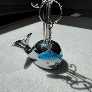 May include: A chrome whale-shaped keychain holder with a silver key ring. A blue beaded fish charm hangs from the key ring. The holder is on a white surface.