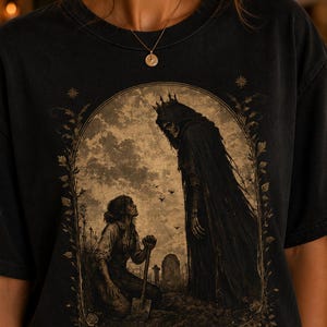 Könnte beinhalten: Schwarzes T-Shirt mit einer gotischen Illustration. Das Design zeigt eine vermummte Gestalt, die einer knienden Person mit einer Schaufel auf einem Friedhof gegenübersteht. Das Kunstwerk ist von floralen Elementen und Sternen umrahmt.