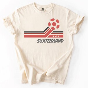 Pode incluir: Uma t-shirt de manga curta na cor creme com um design gr&aacute;fico que apresenta uma bola de futebol vermelha, riscas horizontais pretas e vermelhas, tr&ecirc;s pequenas estrelas brancas e a palavra SWITZERLAND em fonte preta, negrito e sem serifa.