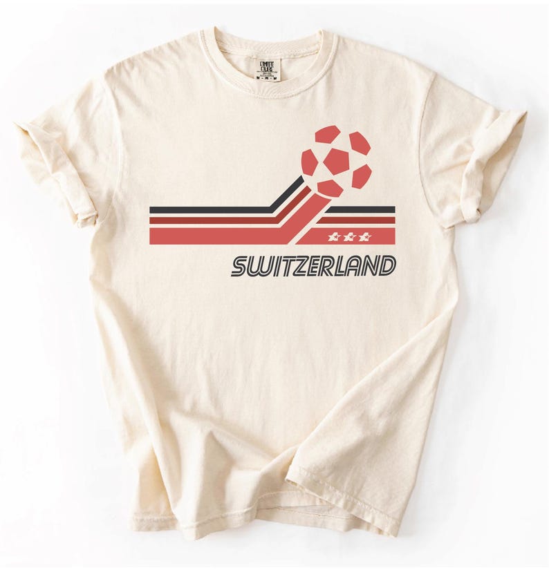 Pode incluir: Uma t-shirt de manga curta na cor creme com um design gr&aacute;fico que apresenta uma bola de futebol vermelha, riscas horizontais pretas e vermelhas, tr&ecirc;s pequenas estrelas brancas e a palavra SWITZERLAND em fonte preta, negrito e sem serifa.