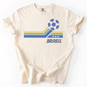 Pode incluir: Uma t-shirt de manga curta na cor creme com um design gr&aacute;fico que apresenta uma bola de futebol azul, riscas horizontais amarelas, verdes e azuis, tr&ecirc;s pequenas estrelas brancas e a palavra BRASIL em letras mai&uacute;sculas azuis.