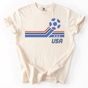 Pode incluir: Uma t-shirt de manga curta na cor creme com um gr&aacute;fico de uma bola de futebol azul, riscas horizontais vermelhas e azuis, tr&ecirc;s pequenas estrelas brancas e o texto USA numa fonte azul contornada na parte da frente da camisola.