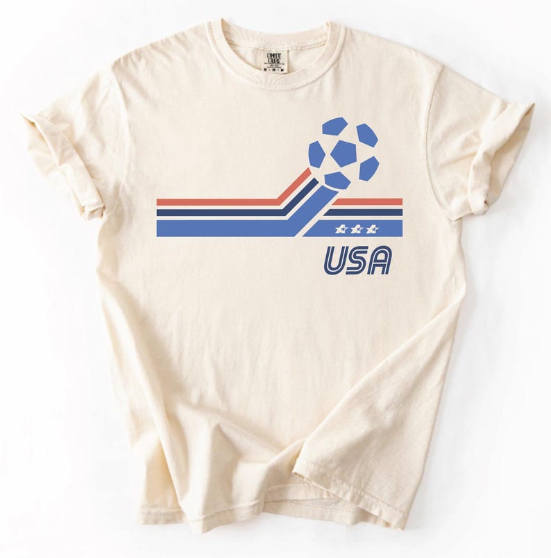 Pode incluir: Uma t-shirt de manga curta na cor creme com um gr&aacute;fico de uma bola de futebol azul, riscas horizontais vermelhas e azuis, tr&ecirc;s pequenas estrelas brancas e o texto USA numa fonte azul contornada na parte da frente da camisola.