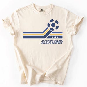 Pode incluir: Uma t-shirt de manga curta cor de creme com um gr&aacute;fico de uma bola de futebol ligada a riscas horizontais azuis e amarelas. Abaixo das riscas, a palavra SCOTLAND est&aacute; impressa em letras azuis, acompanhada por tr&ecirc;s pequenas estrelas brancas.