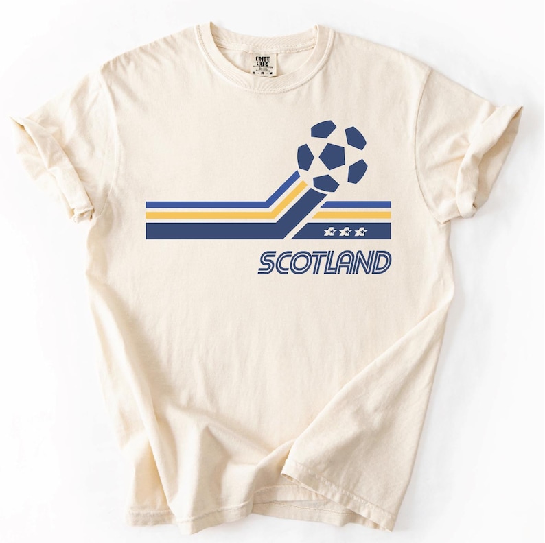 Pode incluir: Uma t-shirt de manga curta cor de creme com um gr&aacute;fico de uma bola de futebol ligada a riscas horizontais azuis e amarelas. Abaixo das riscas, a palavra SCOTLAND est&aacute; impressa em letras azuis, acompanhada por tr&ecirc;s pequenas estrelas brancas.
