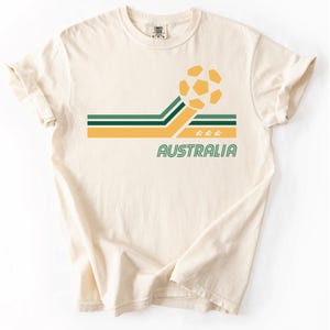 Pode incluir: Uma t-shirt de manga curta cor de creme com um gr&aacute;fico de uma bola de futebol amarela, riscas horizontais verdes e amarelas, a palavra AUSTRALIA impressa em verde e tr&ecirc;s pequenos &iacute;cones de estrelas amarelas sob as riscas.