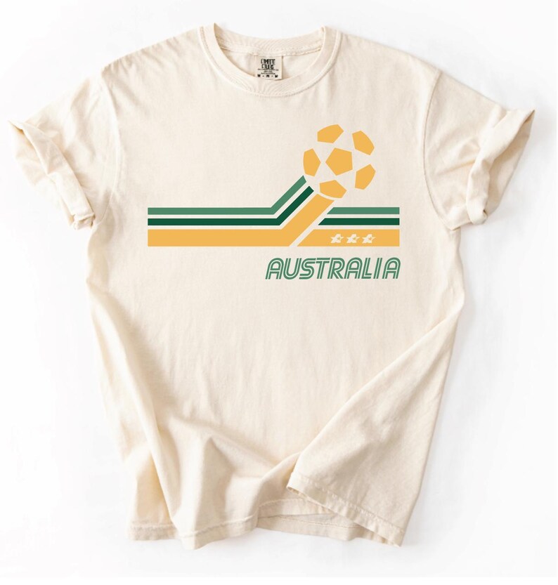 Pode incluir: Uma t-shirt de manga curta cor de creme com um gr&aacute;fico de uma bola de futebol amarela, riscas horizontais verdes e amarelas, a palavra AUSTRALIA impressa em verde e tr&ecirc;s pequenos &iacute;cones de estrelas amarelas sob as riscas.