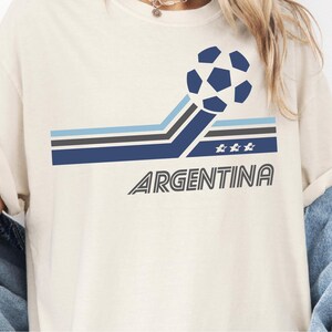 Pode incluir: Uma t-shirt de cor creme com um gr&aacute;fico de uma bola de futebol azul-escura com riscas angulares em azul-claro, cinzento e azul-escuro. O design inclui tr&ecirc;s pequenas estrelas brancas e a palavra ARGENTINA numa fonte sem serifa azul-escura.