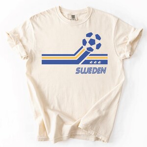 Pode incluir: Uma t-shirt de manga curta na cor creme com um design gr&aacute;fico que apresenta uma bola de futebol azul e amarela, riscas horizontais, tr&ecirc;s pequenas estrelas e a palavra SWEDEN impressa em letras mai&uacute;sculas azuis no centro do peito.