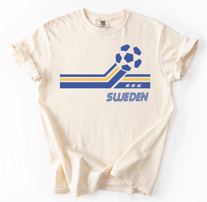 Pode incluir: Uma t-shirt de manga curta na cor creme com um design gr&aacute;fico que apresenta uma bola de futebol azul e amarela, riscas horizontais, tr&ecirc;s pequenas estrelas e a palavra SWEDEN impressa em letras mai&uacute;sculas azuis no centro do peito.