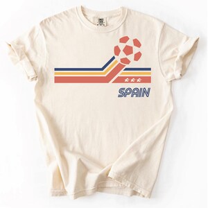 Pode incluir: Uma t-shirt de manga curta na cor creme com um gr&aacute;fico de uma bola de futebol e riscas horizontais em vermelho, amarelo e azul. O design inclui tr&ecirc;s pequenas estrelas e a palavra SPAIN impressa em letras mai&uacute;sculas azuis abaixo.
