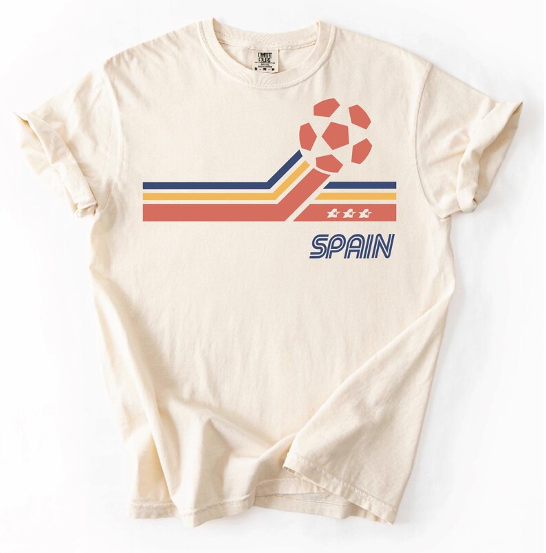 Pode incluir: Uma t-shirt de manga curta na cor creme com um gr&aacute;fico de uma bola de futebol e riscas horizontais em vermelho, amarelo e azul. O design inclui tr&ecirc;s pequenas estrelas e a palavra SPAIN impressa em letras mai&uacute;sculas azuis abaixo.