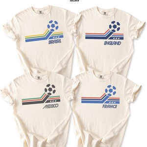 Pode incluir: Quatro t-shirts de cor marfim Comfort Colors 1717 com um gr&aacute;fico de bola de futebol, riscas coloridas e o texto Brasil, England, Mexico e France. Cada t-shirt apresenta um esquema de cores nacional &uacute;nico e tr&ecirc;s estrelas sob a bola.