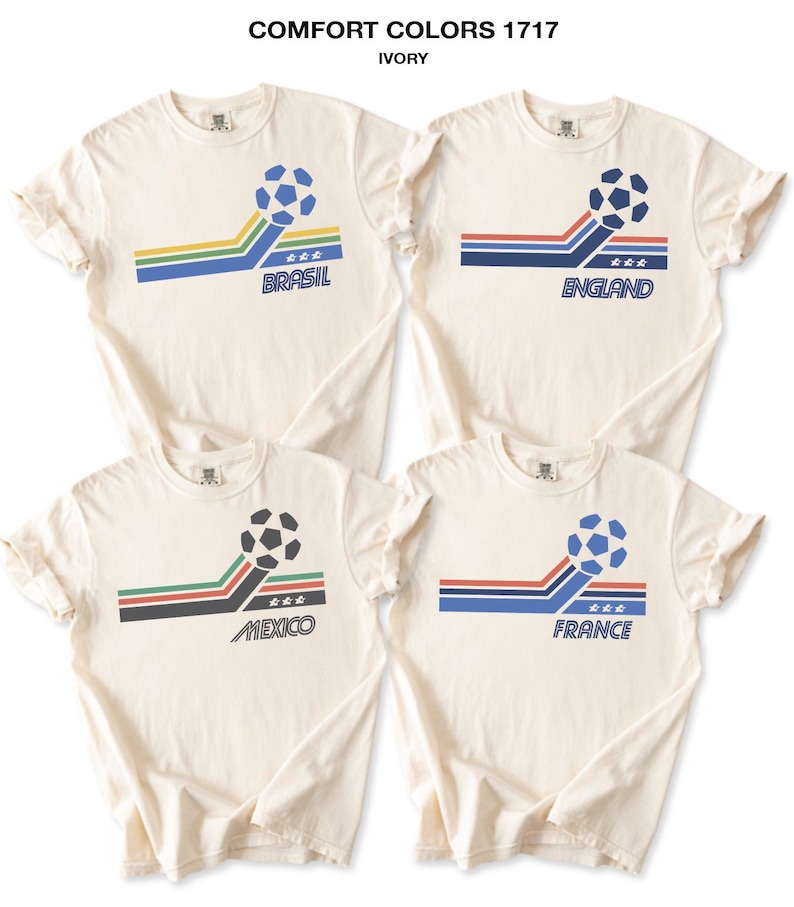 Pode incluir: Quatro t-shirts de cor marfim Comfort Colors 1717 com um gr&aacute;fico de bola de futebol, riscas coloridas e o texto Brasil, England, Mexico e France. Cada t-shirt apresenta um esquema de cores nacional &uacute;nico e tr&ecirc;s estrelas sob a bola.