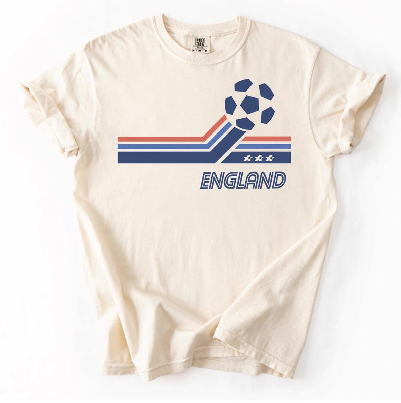 Pode incluir: Uma t-shirt de manga curta cor de creme com um gr&aacute;fico de uma bola de futebol sobre riscas horizontais vermelhas, brancas e azuis. O design inclui tr&ecirc;s pequenas estrelas e a palavra "ENGLAND" em letras azuis contornadas sob as riscas.