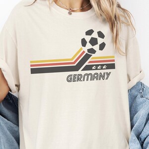Pode incluir: Uma t-shirt de cor creme com um gr&aacute;fico de uma bola de futebol sobre tr&ecirc;s faixas horizontais em amarelo, vermelho e preto. As faixas inclinam-se para cima em dire&ccedil;&atilde;o &agrave; bola. Abaixo das faixas, a palavra GERMANY est&aacute; impressa com tr&ecirc;s estrelas.
