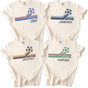 Pode incluir: Quatro t-shirts de manga curta em branco sujo com um gr&aacute;fico de bola de futebol, riscas inclinadas e nomes de pa&iacute;ses. As t-shirts exibem o texto USA, ARGENTINA, GERMANY e PORTUGAL, cada uma acompanhada por tr&ecirc;s pequenas estrelas e riscas coloridas.