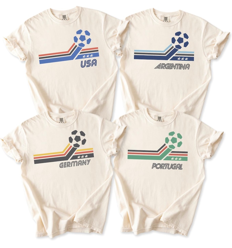 Pode incluir: Quatro t-shirts de manga curta em branco sujo com um gr&aacute;fico de bola de futebol, riscas inclinadas e nomes de pa&iacute;ses. As t-shirts exibem o texto USA, ARGENTINA, GERMANY e PORTUGAL, cada uma acompanhada por tr&ecirc;s pequenas estrelas e riscas coloridas.
