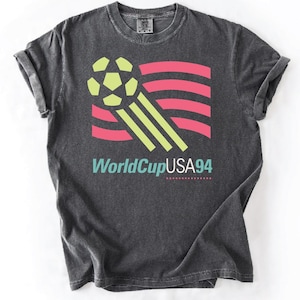 May include: Dark gray t-shirt featuring a soccer ball and a flag design in pink, green, and yellow. The text "WorldCupUSA94" is printed below the graphic in teal and white. The shirt has rolled-up sleeves.