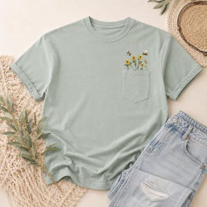 May include: A sage green short-sleeve t-shirt featuring a chest pocket with a printed graphic of yellow sunflowers and small bees. The shirt has a crew neckline and a relaxed fit, showcasing a botanical design on the pocket area.