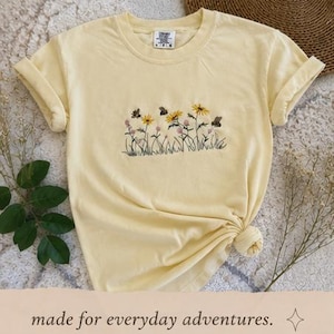 May include: A pale yellow t-shirt featuring embroidered wildflowers and butterflies. The sleeves are rolled up, and the hem is knotted. The text "made for everyday adventures" is visible at the bottom of the image.