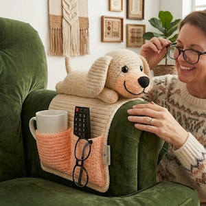 May include: A beige crochet dog-shaped armrest cover with a peach-colored pocket for a mug, remote, and glasses. The dog has a black nose and smiling face. The armrest cover is on a green armchair.