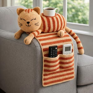May include: A crocheted cat-shaped armrest organizer in shades of orange and cream, holding a white mug of coffee. The organizer has pockets for remote controls and a striped pattern.
