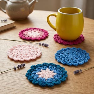 May include: A selection of colourful, handmade crochet coasters in shades of pink and blue, arranged on a wooden table. The coasters are round with a scalloped edge. A yellow mug and a teapot are also visible in the image.
