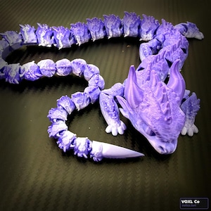 May include: A purple articulated dragon figurine. The dragon is 3D-printed and has a segmented body, head, and limbs. The dragon's head features horns and a detailed face. The dragon is lying on a dark surface.