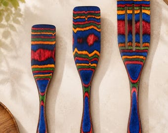 May include: A set of three kitchen utensils made of multi-colored laminated wood featuring horizontal stripes in blue, red, green, and orange. The set includes two solid spatulas of varying sizes and one slotted spatula, each with a hole in the handle.