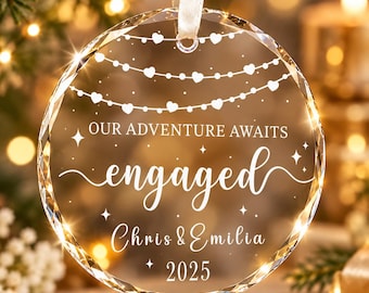 May include: A circular clear glass ornament with beveled edges, hanging from a white ribbon. The surface features white etched text reading "OUR ADVENTURE AWAITS", "engaged", "Chris & EMILIA", and "2025", accented by heart-shaped string lights and stars.
