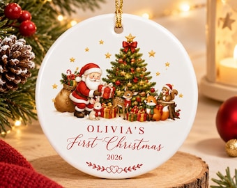 May include: A round white ceramic Christmas ornament featuring a colorful illustration of Santa Claus, a deer, a fox, a squirrel, and a rabbit around a decorated tree. The text reads OLIVIA'S First Christmas 2026 with red heart accents.