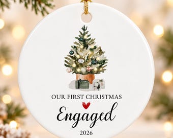 May include: A round white ceramic ornament featuring a watercolor illustration of a decorated Christmas tree with gifts underneath. The text reads "OUR FIRST CHRISTMAS" above a red heart and the word "Engaged" in script, with "2026" below.