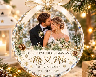May include: A circular glass Christmas ornament featuring a photograph of a couple in wedding attire. The ornament is decorated with gold pinecones, greenery, and snowflakes. Text reads: OUR FIRST CHRISTMAS AS Mr & Mrs EMILY & JAMES 2026.
