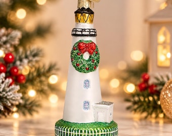 May include: A glass lighthouse ornament featuring a white tower with a black lantern room, a green base with a white fence pattern, and a green wreath with a red bow attached to the front. A gold heart-shaped charm hangs from the top loop.