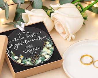 May include: A round black acrylic ornament with white script text reading the best is yet to come, a line drawing of two rings, and the word ENGAGED 2026, decorated with green eucalyptus leaves and gold stems, presented in a brown cardboard gift box.
