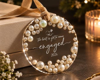 May include: A circular clear acrylic ornament decorated with white pearls of varying sizes along the edge. The center features an engraved graphic of two rings and the text: the best is yet to come, engaged, 2026. A beige velvet ribbon is attached to the top.