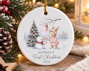 May include: A round white ceramic ornament featuring a printed illustration of a rabbit in a pink sweater building a snowman near a Christmas tree. A red stocking hangs from a branch above. The text reads SOPHIA'S First Christmas 2026.