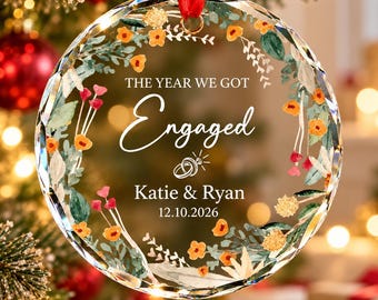 May include: A circular clear glass ornament with faceted edges, featuring a watercolor floral wreath design in orange, red, and green. The text reads THE YEAR WE GOT Engaged, Katie & Ryan, and 12.10.2026, with a small ring icon in the center.