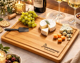 May include: A rectangular bamboo serving board featuring a juice groove and the engraved text The Zimmermans ESTABLISHED 2022. The board holds a wedge of brie with a rosemary sprig, blue cheese, and cheddar cubes, accompanied by a black cheese spreader.