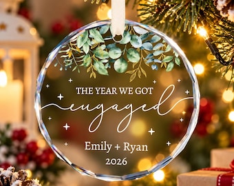 May include: A circular clear glass ornament with faceted edges, featuring a white printed design of eucalyptus leaves, the text THE YEAR WE GOT engaged, the names Emily + Ryan, and the year 2026, suspended by a white ribbon.