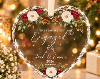May include: A heart-shaped clear glass ornament featuring a floral design with white flowers, red berries, and green leaves. The text reads THE YEAR WE GOT Engaged, Jack & Emma, and 2026, with icons of two interlocking rings and two clinking champagne flutes.