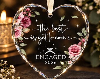 May include: A heart-shaped clear glass ornament with faceted edges, featuring a cream ribbon hanger. The design includes pink roses, green leaves, and the text "the best is yet to come", "ENGAGED", and "2026" with a diamond ring icon.