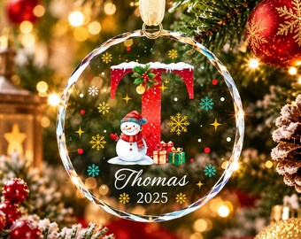 May include: A circular clear glass Christmas ornament featuring a large red letter T topped with snow, a snowman wearing a blue hat and red scarf, and colorful gift boxes. The text Thomas and 2025 are printed below, surrounded by gold snowflakes.