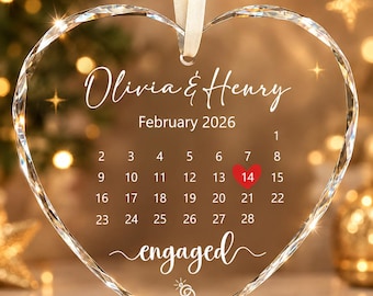 May include: A heart-shaped clear glass ornament with faceted edges, hanging from a white ribbon. It features the names Olivia & Henry, the date February 2026, a calendar with the 14th marked by a red heart, the word engaged, and a ring icon.