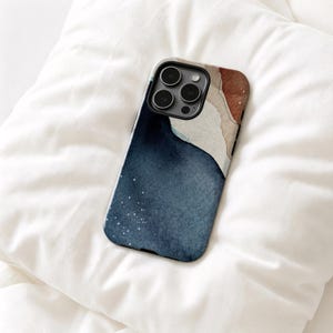May include: A smartphone case featuring an abstract watercolor design with a deep navy blue section, a textured beige area, and a warm terracotta accent. The case has a cutout for a triple-lens camera system and small white speckles.