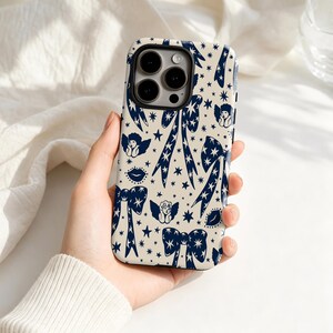 May include: A smartphone case with a cream-colored background featuring a repeating dark blue pattern of bows, small stars, cherubs, and lips. The case has a cutout for a triple-lens camera system on the upper left side and black edges.