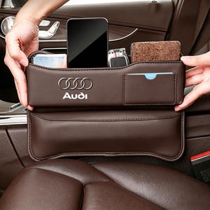 May include: A brown leather car seat gap organizer featuring the Audi logo and text. The organizer has multiple compartments holding a smartphone, a key fob, a brown wallet, and a small card, designed to fit between the seat and center console.