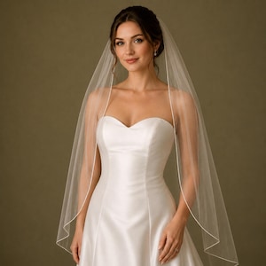 May include: A bride in a white strapless wedding dress and a sheer white veil with a delicate white border. The bride is smiling. The dress has a sweetheart neckline.